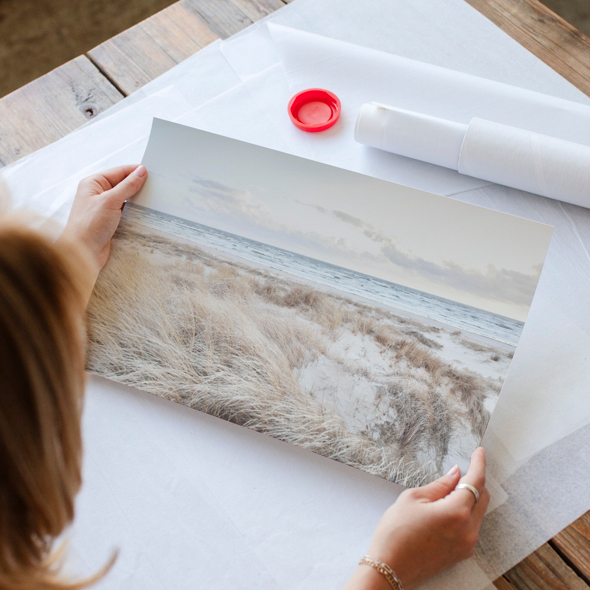 A person is holding a framed photograph of a beach scene, with a red bottle and a roll of paper visible on the table.