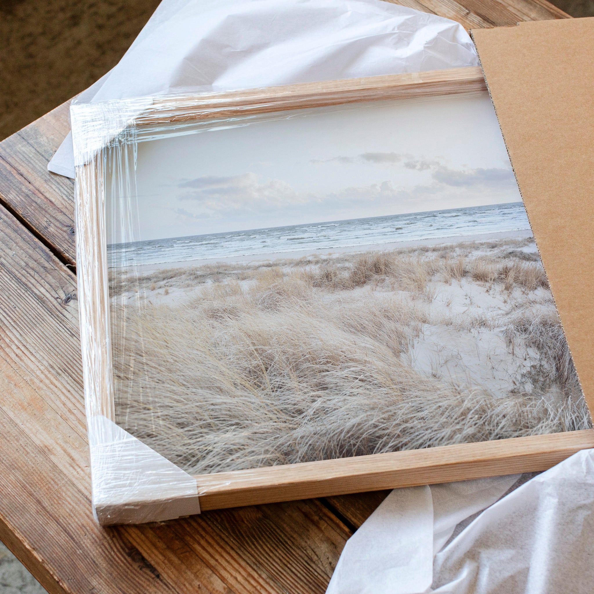 A framed photograph of a beach scene with a cloudy sky and grassy landscape is displayed on a wooden surface.