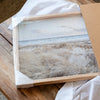 A framed photograph of a beach scene with a cloudy sky and grassy landscape is displayed on a wooden surface.