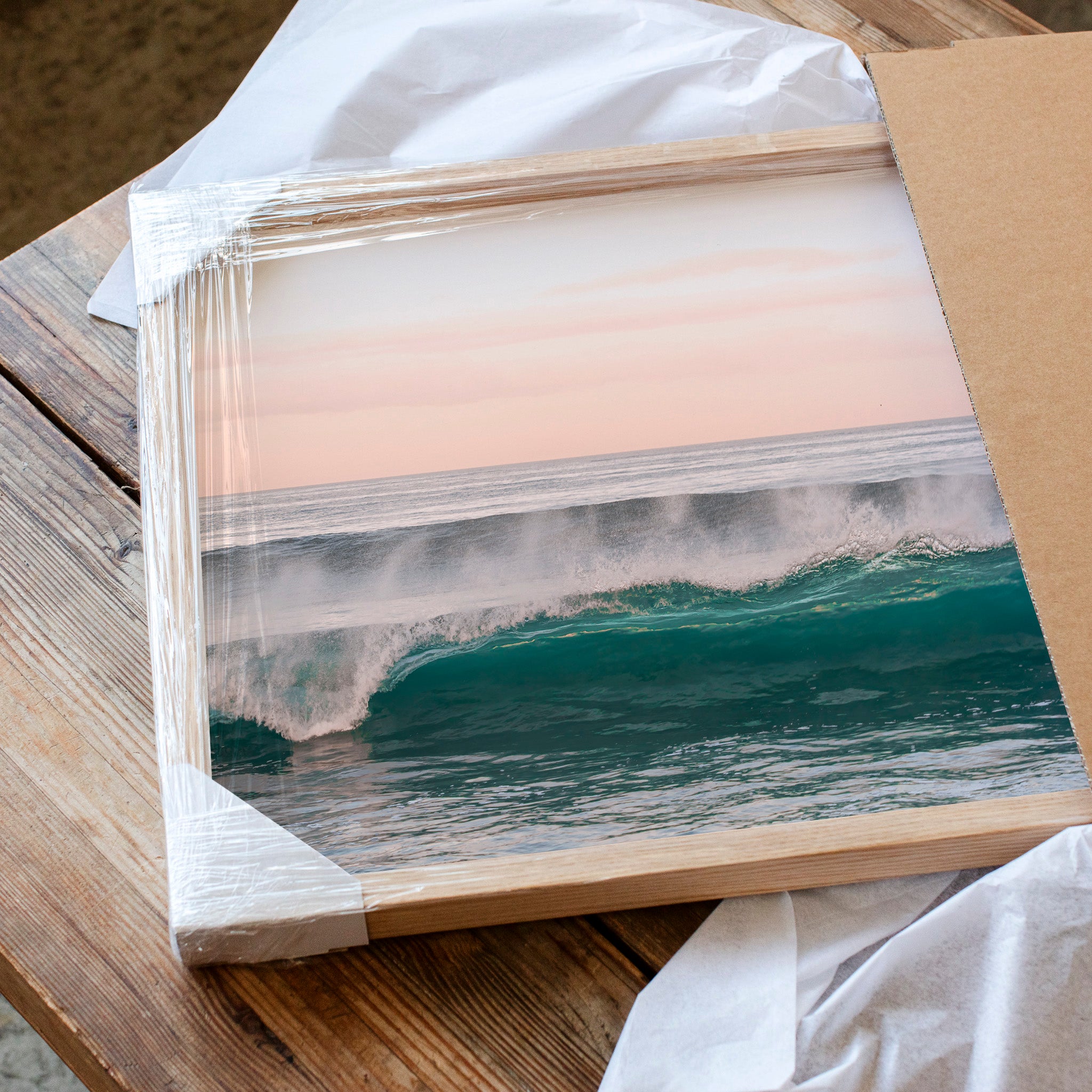 A framed photograph of a crashing wave is displayed on a wooden surface, with a piece of cardboard visible in the background.