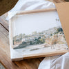 a framed photograph of a beach scene, with a wooden frame and a clear plastic cover. The photograph appears to be a collage of multiple images, and the image is not described in detail.