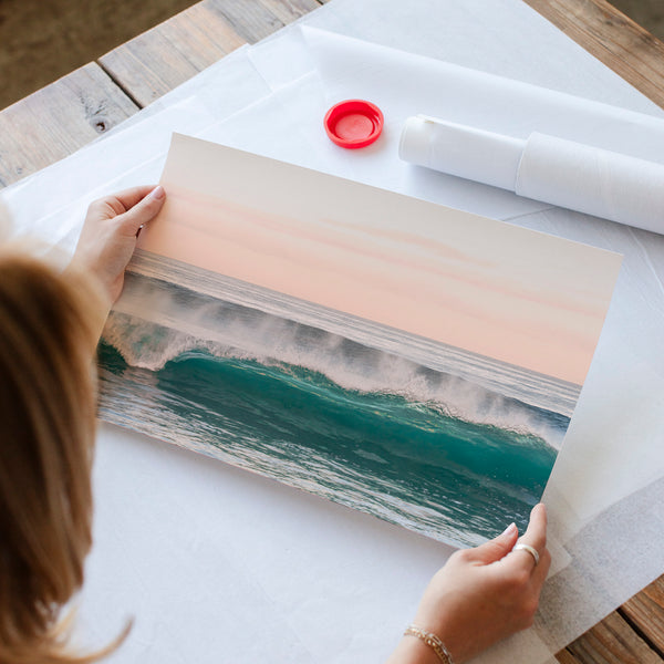 A person is holding a framed print of a seascape with crashing waves, and there is a red bottle cap on the table.