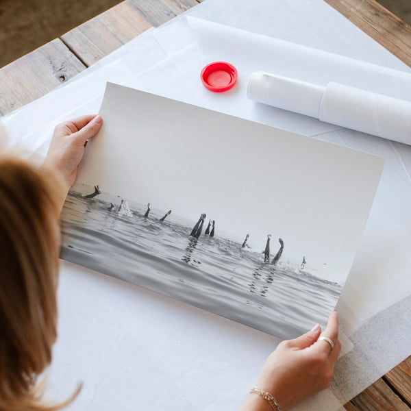 A person is holding a framed black and white photograph of a beach scene, with a red bottle and a roll of paper towels visible on the table.