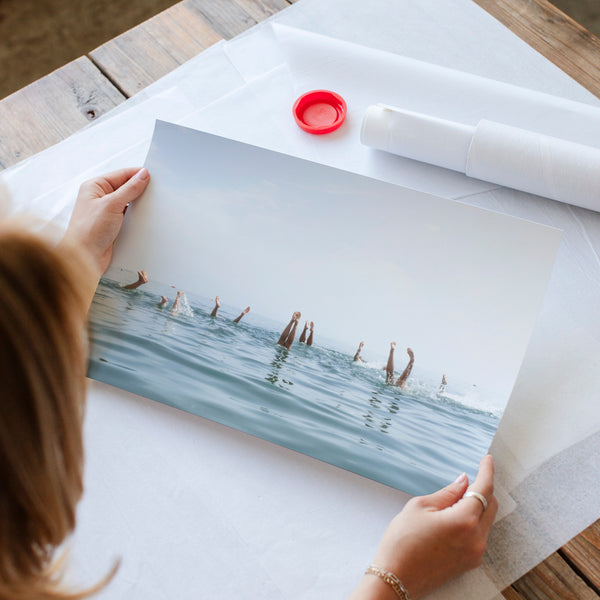 A person is holding a framed photograph of a group of people swimming in the ocean, with the ocean waves visible in the background.