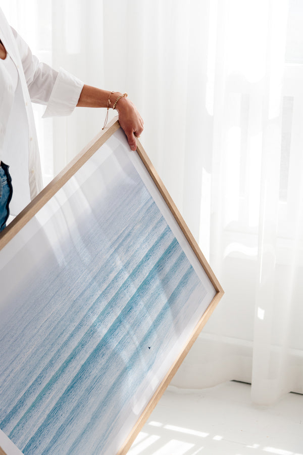 Woman holding a framed art print in a sunlit room featuring a coastal surf photography beach scene.