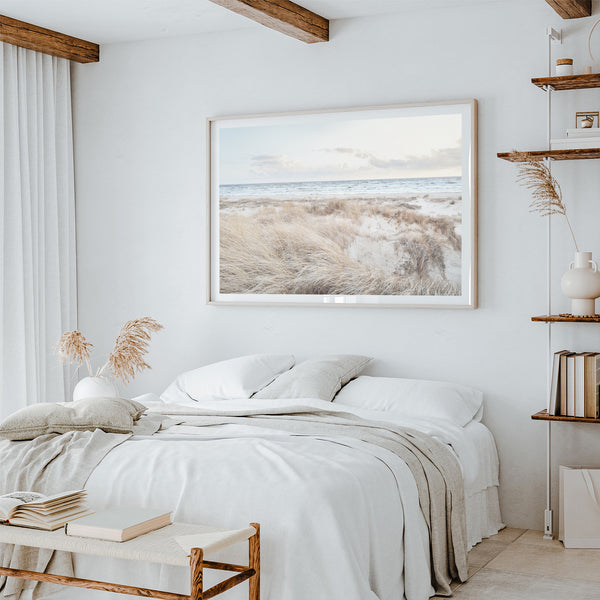 a serene bedroom with a large bed, a framed picture of a beach scene, and a bookshelf filled with books.